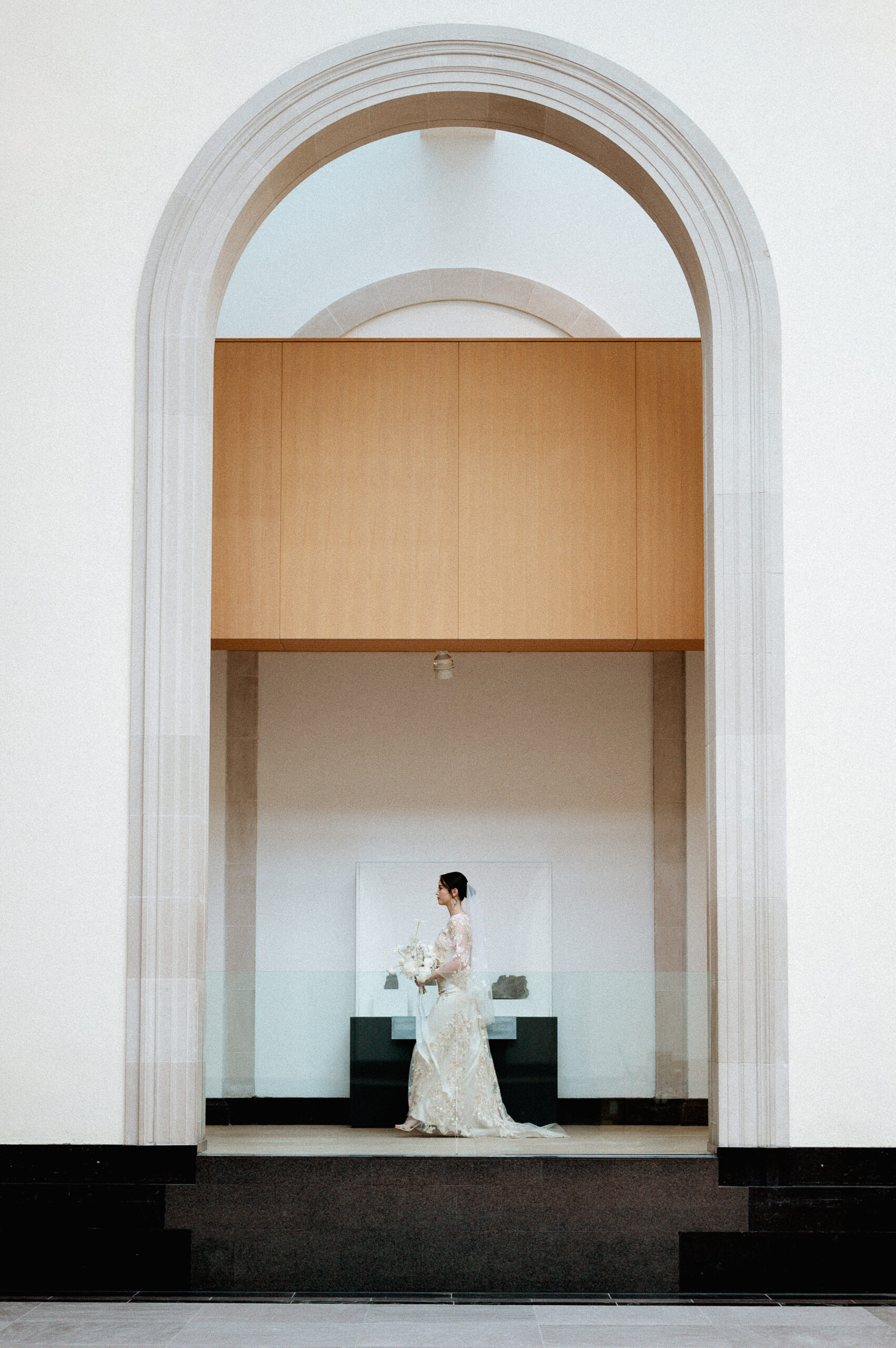 "A bride in an elegant lace wedding gown walking gracefully through a grand arched hallway, holding a white bouquet. The setting features minimalist architecture with clean lines and a modern, serene atmosphere.