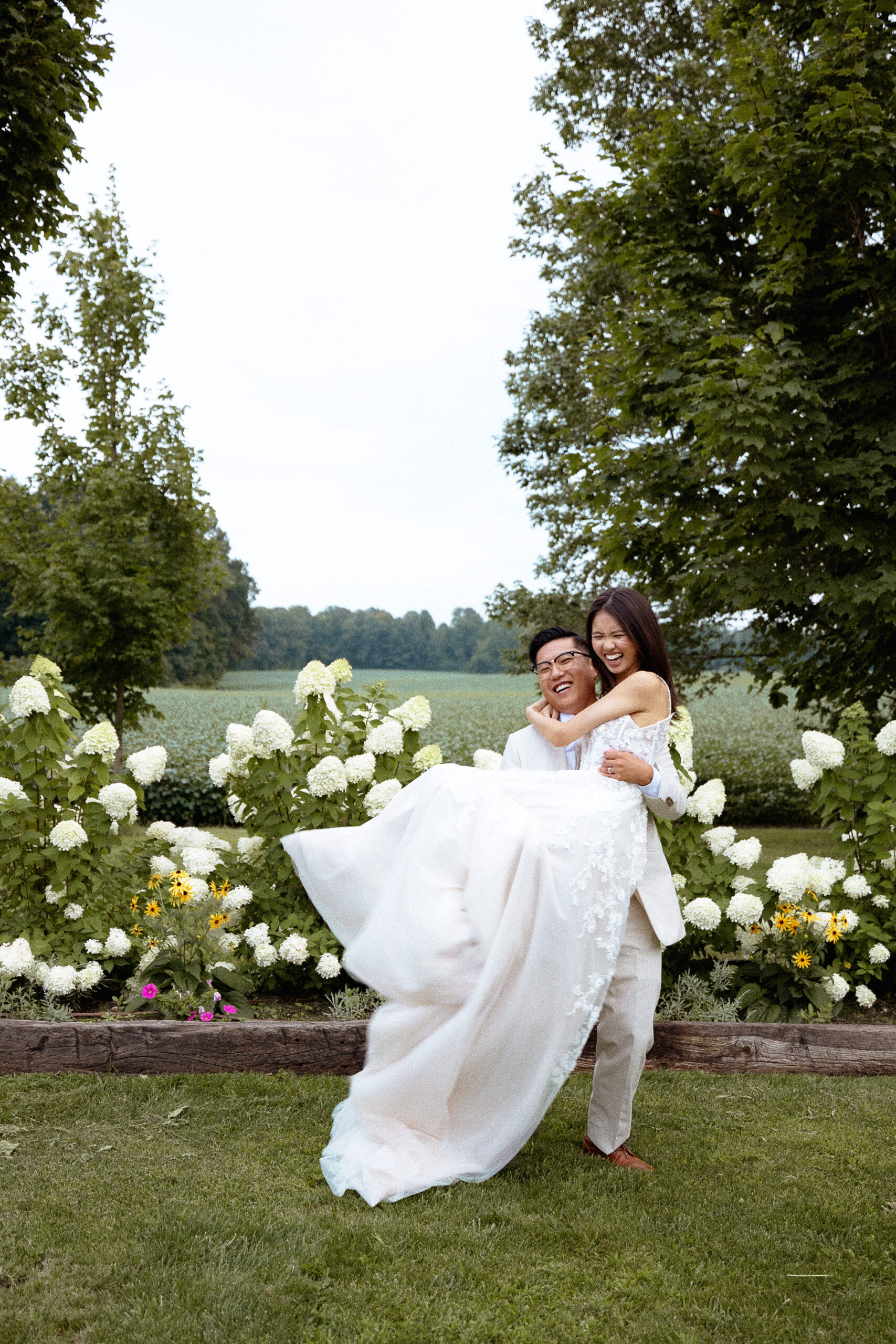 A joyful bride and groom sharing a playful moment in a garden, with the groom lifting the bride in front of blooming flowers, capturing a candid and lighthearted wedding portrait.