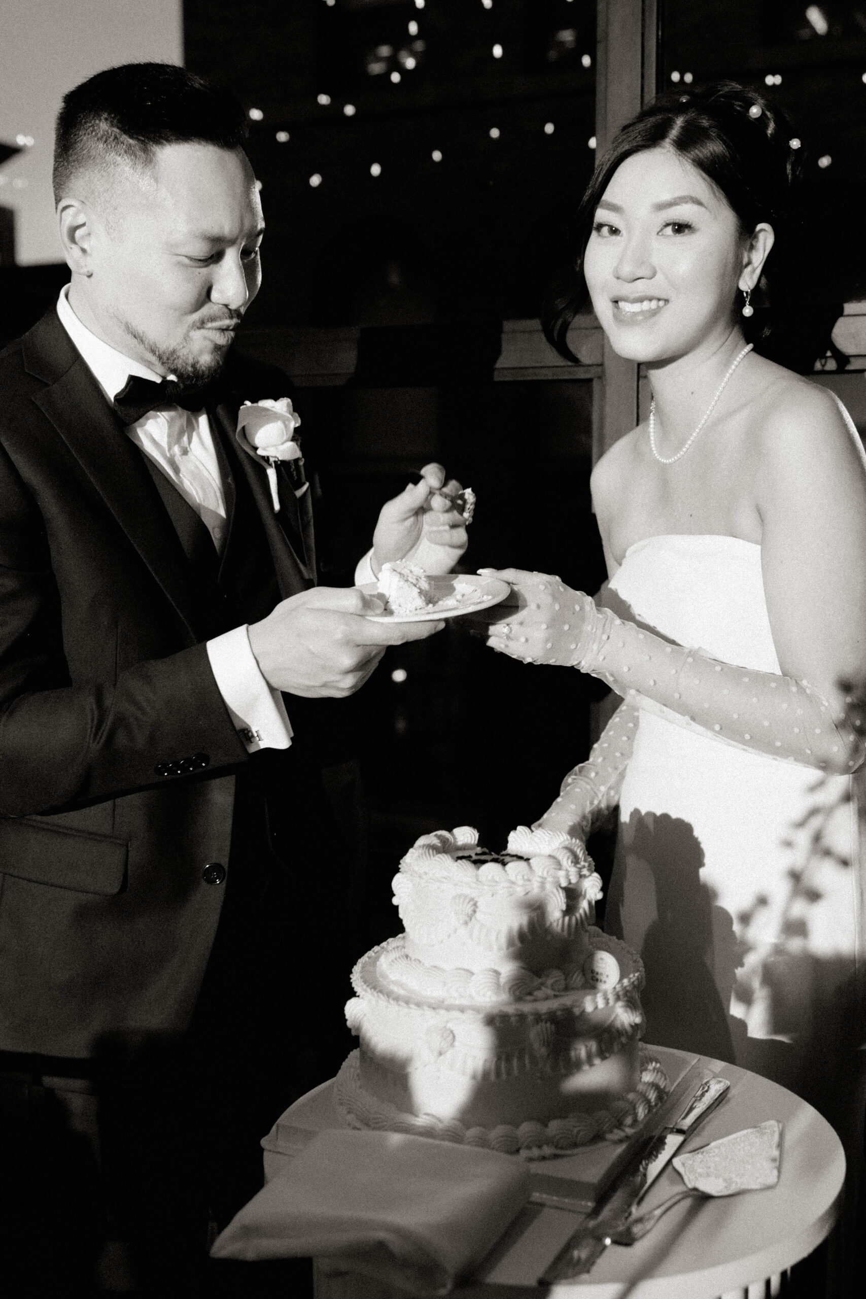 A bride and groom share a lighthearted moment as they cut their wedding cake together. The bride smiles at the camera while the groom playfully prepares a bite of cake. The scene is lit with romantic string lights in the background.