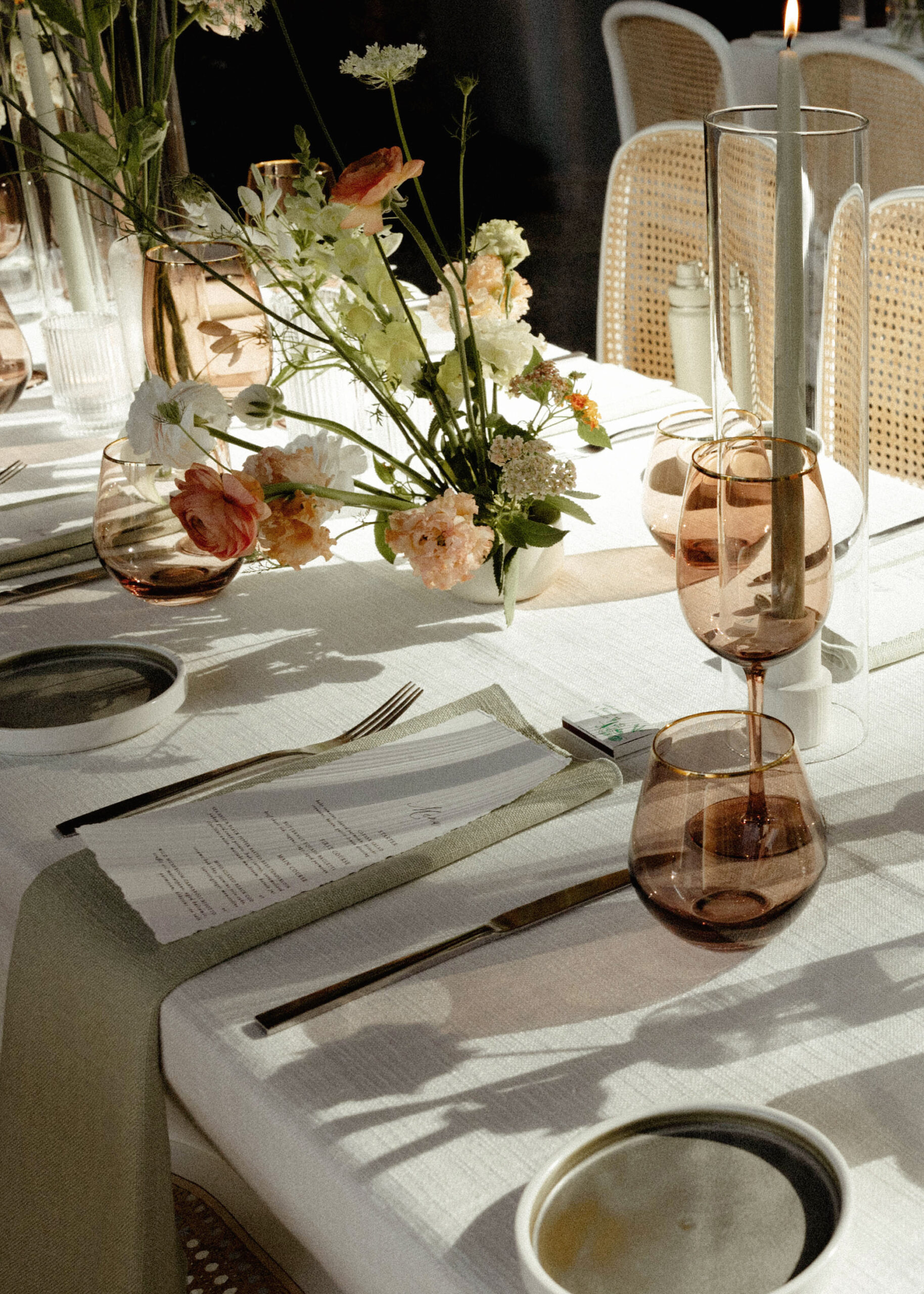 A luxuriously styled wedding reception table bathed in natural light, featuring elegant floral centerpieces with soft peach and white blooms. The minimalist yet refined setup includes amber-tinted wine glasses, delicate candle holders, and menus placed atop olive-hued napkins on crisp white linens. The overall aesthetic is warm, intimate, and sophisticated, with shadows cast across the table adding depth to the serene atmosphere. Cane-back chairs surround the table, enhancing the organic, high-end feel perfect for a modern, upscale wedding celebration.