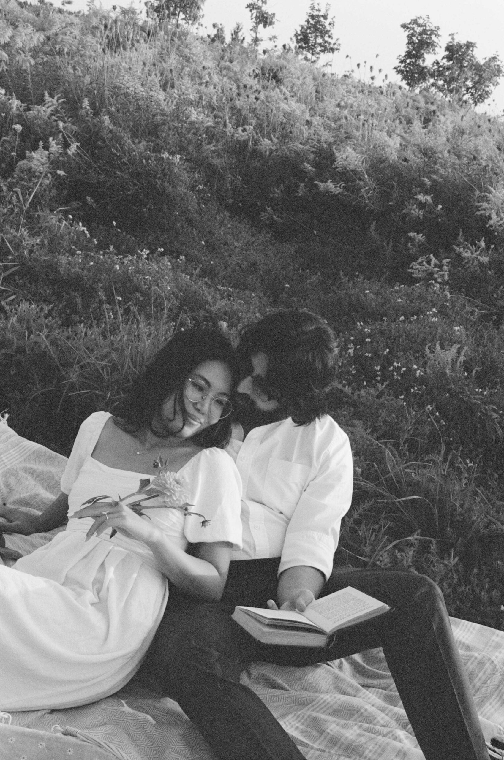 Black and white photo of a couple relaxing on a picnic blanket in a meadow. The woman is leaning back with a soft smile, holding a flower, while the man sits close behind her, reading a book and gently resting his head against hers. The scene feels intimate and peaceful, surrounded by wildflowers and natural greenery.