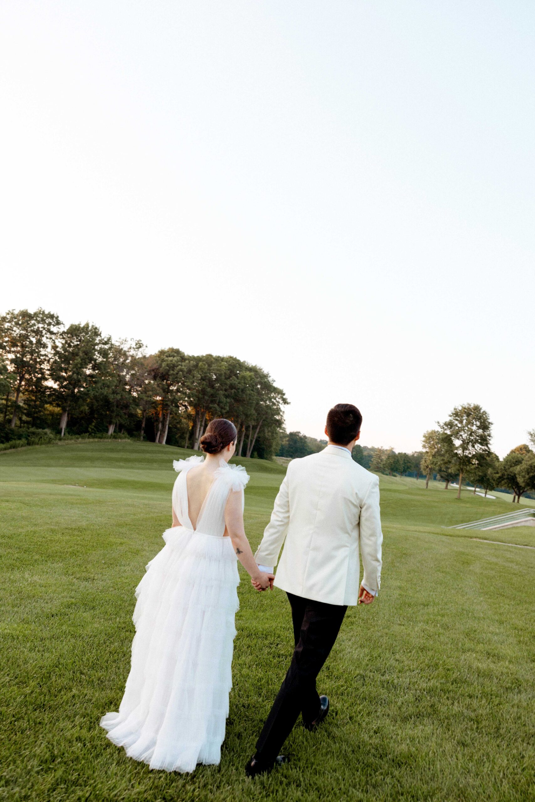 A bride and groom walk hand-in-hand across a grassy landscape, backs turned toward the camera. The bride wears a white gown with delicate tulle and shoulder details, her hair elegantly styled in a low bun. The groom is dressed in a white tuxedo jacket and black pants. Trees line the background under a clear sky, creating a serene and timeless setting for the couple's wedding moment.