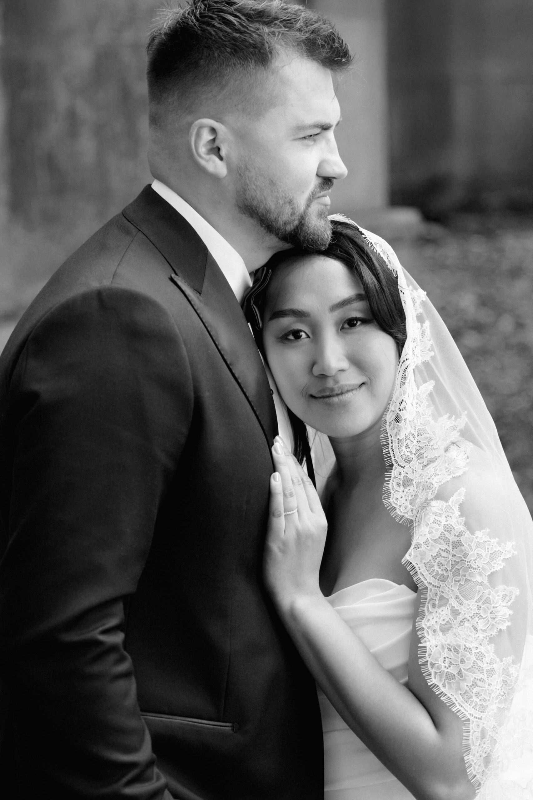 Black-and-white portrait of a bride and groom embracing outdoors; the groom in a suit looks off into the distance while the bride, wearing a lace-trimmed veil and strapless gown, rests her head against him and looks toward the camera with a soft, calm expression.