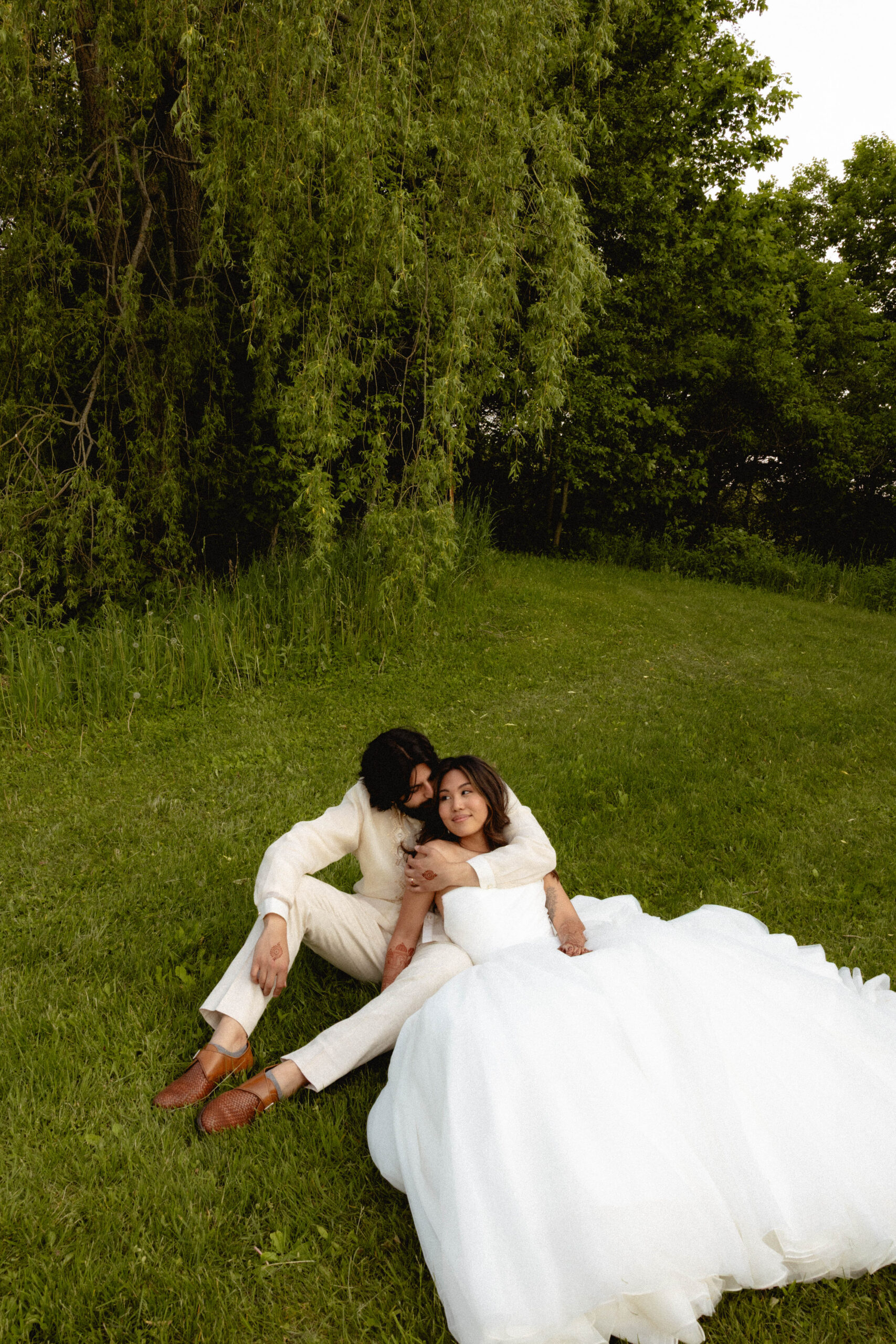 Bride and groom lying together on green grass at Mount Alverno, the bride in a white tulle gown and the groom in a cream suit, sharing a quiet, intimate moment beneath a willow tree during their wedding day.