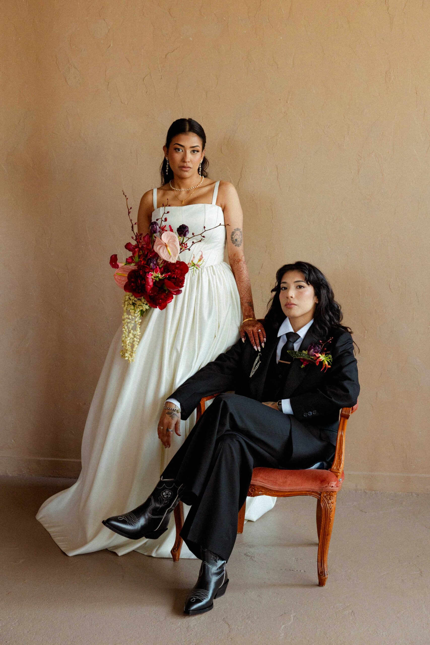 Gabriele and Zai in an editorial portrait at their Toronto city wedding, one standing in an ivory gown with a red bouquet, one seated in a black suit and boots.
