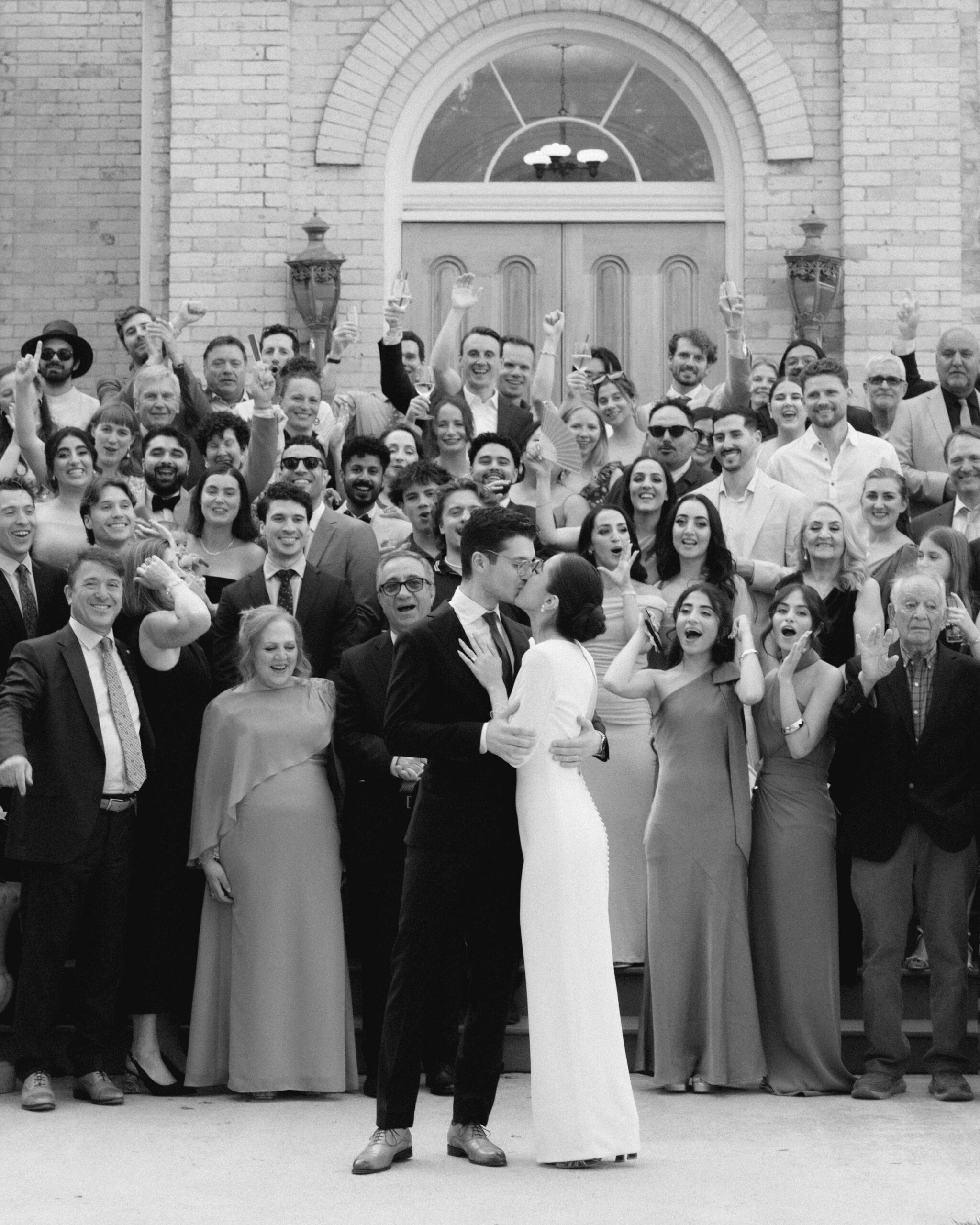 Madina and Olivier kissing in front of their cheering guests at their Afghan fusion wedding in Toronto, the full group gathered on the steps behind them.